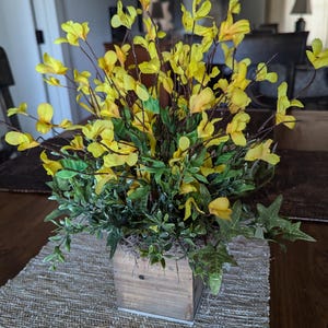 May include: A wooden planter box filled with a floral arrangement of yellow flowers and green leaves.