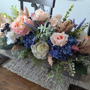 May include: A floral centerpiece in a wooden box with pink, white, and blue flowers, including roses and hydrangeas. The centerpiece is surrounded by greenery and dried grasses.
