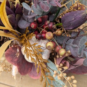 May include: A close-up of a fall-themed floral arrangement. The arrangement features deep purple, burgundy, and gold-toned foliage, including berries, eucalyptus, and pinecones. The colors create a warm, autumnal aesthetic.