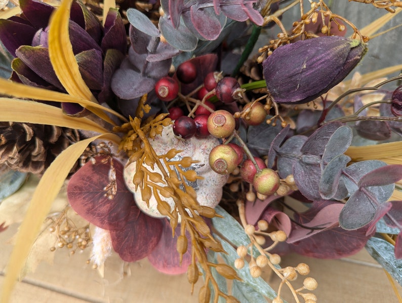 May include: A close-up of a fall-themed floral arrangement. The arrangement features deep purple, burgundy, and gold-toned foliage, including berries, eucalyptus, and pinecones. The colors create a warm, autumnal aesthetic.