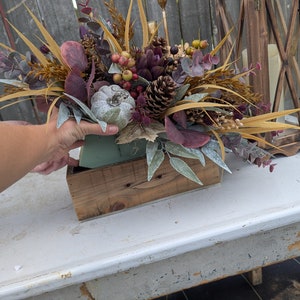 May include: A fall-themed floral arrangement in a rustic wooden box. The arrangement features a variety of textures and colors, including faux pumpkins, pinecones, berries, and dried foliage. The color palette includes shades of brown, green, purple, and gold.