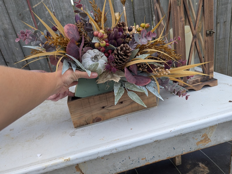 May include: A fall-themed floral arrangement in a rustic wooden box. The arrangement features a variety of textures and colors, including faux pumpkins, pinecones, berries, and dried foliage. The color palette includes shades of brown, green, purple, and gold.