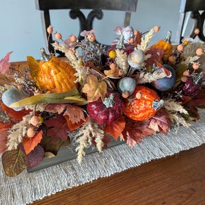 May include: A fall-themed centerpiece featuring a wooden box filled with decorative pumpkins, berries, and leaves in shades of orange, red, and brown. The arrangement includes faux pumpkins and various dried foliage.