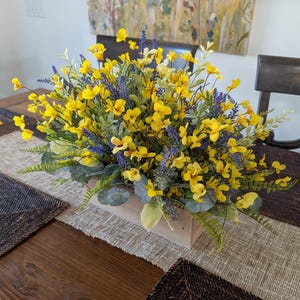 May include: A wooden rectangular centerpiece filled with vibrant yellow flowers and purple accents. Green foliage and leaves surround the flowers, creating a natural and colorful arrangement. The centerpiece sits on a neutral-toned table runner.