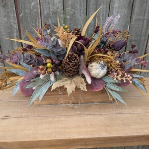 May include: A fall-themed floral arrangement in a wooden box. The arrangement features a variety of dried flowers, including eucalyptus, berries, and wheat. The color palette includes shades of purple, gold, and green. A small decorative pumpkin and pinecone are also included.