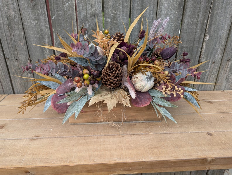 May include: A fall-themed floral arrangement in a wooden box. The arrangement features a variety of dried flowers, including eucalyptus, berries, and wheat. The color palette includes shades of purple, gold, and green. A small decorative pumpkin and pinecone are also included.