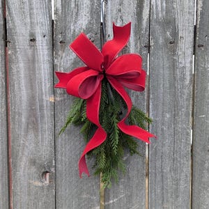 May include: A red velvet bow tied around a sprig of evergreen, hanging on a weathered wooden fence.