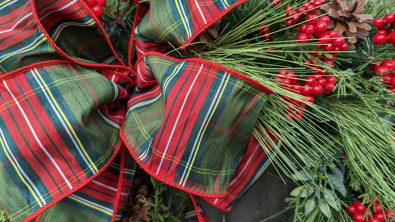 May include: A green and red plaid ribbon with a red trim, tied into a bow. The ribbon is decorated with sprigs of green pine and red berries.