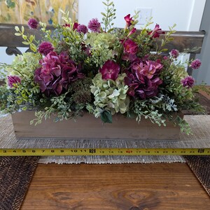 Pu&ograve; includere: Una fioriera rettangolare in legno riempita con una composizione di fiori finti. La composizione comprende ortensie viola intenso, ortensie verdi e piccoli fiori globulari viola. La scatola &egrave; lunga circa 61 cm.