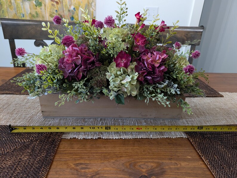 Pu&ograve; includere: Una fioriera rettangolare in legno riempita con una composizione di fiori finti. La composizione comprende ortensie viola intenso, ortensie verdi e piccoli fiori globulari viola. La scatola &egrave; lunga circa 61 cm.