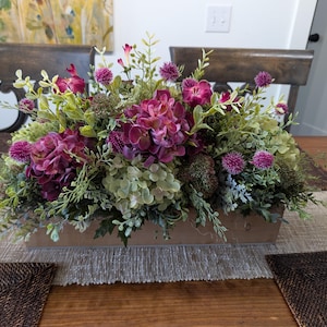 Pu&ograve; includere: Un'aiuola rettangolare in legno con una vivace composizione di fiori artificiali. La composizione presenta ortensie viola scuro, fiori rosa pi&ugrave; piccoli e varie foglie verdi. L'aiuola &egrave; posizionata su un runner da tavolo dai toni neutri.