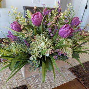 May include: A floral arrangement featuring vibrant purple tulips, green hydrangeas, and delicate lavender sprigs. The flowers are arranged in a rectangular wooden box, creating a fresh and colorful centerpiece for a table setting.