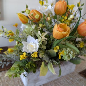 May include: A floral arrangement featuring orange tulips, white daffodils, and yellow blossoms in a rectangular, whitewashed wooden container. Green foliage and accents complete the spring-themed display.