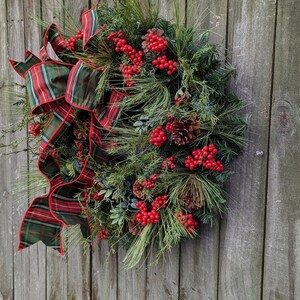 May include: A green and red plaid ribbon bow decorates a Christmas wreath with pine branches, red berries, and pine cones.