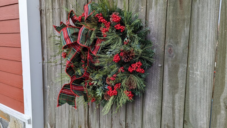 May include: A green and red plaid ribbon bow decorates a Christmas wreath with pine branches, red berries, and pine cones.