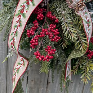 May include: A festive Christmas wreath with evergreen branches, red berries, and a decorative ribbon. The ribbon has a beige background with a red border and a pattern of green pine needles and red berries. A pine cone is also visible.