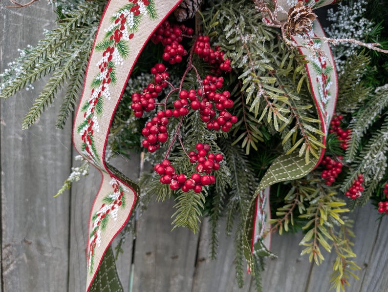 May include: A festive Christmas wreath with evergreen branches, red berries, and a decorative ribbon. The ribbon has a beige background with a red border and a pattern of green pine needles and red berries. A pine cone is also visible.