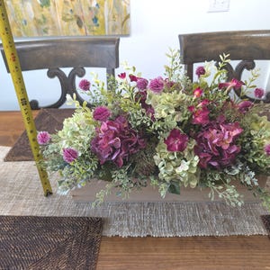 Pu&ograve; includere: Un'arrangiamento floreale artificiale in una fioriera rettangolare in legno. Include ortensie viola, ortensie verdi e bianche e piccoli fiori rosa. L'arrangiamento &egrave; lungo circa 30 cm.