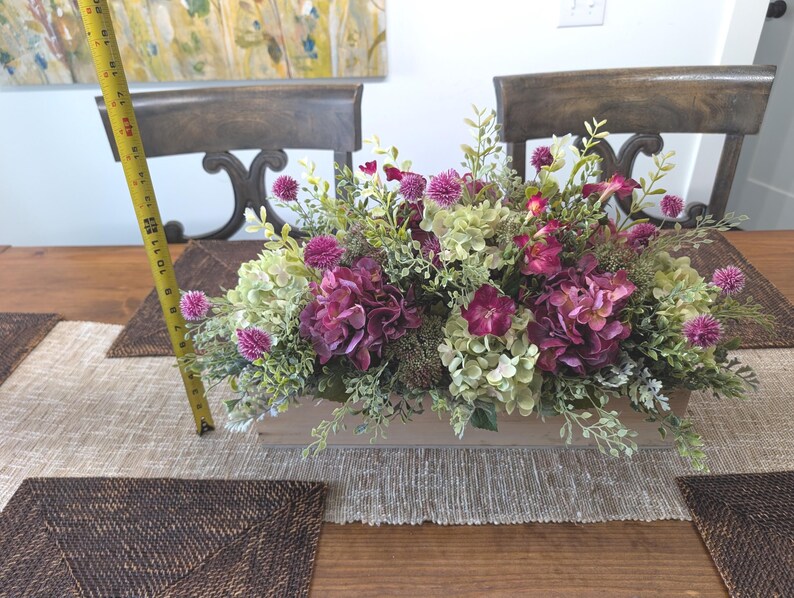 Pu&ograve; includere: Un'arrangiamento floreale artificiale in una fioriera rettangolare in legno. Include ortensie viola, ortensie verdi e bianche e piccoli fiori rosa. L'arrangiamento &egrave; lungo circa 30 cm.