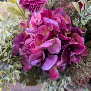 Pu&ograve; includere: Primo piano di una vivace composizione floreale con un grande gruppo di fiori di ortensia magenta intenso. Intorno alle ortensie, fiori viola pi&ugrave; piccoli e appuntiti e vari accenti di foglie verdi. La composizione &egrave; lussureggiante e colorata.