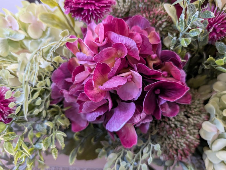 Pu&ograve; includere: Primo piano di una vivace composizione floreale con un grande gruppo di fiori di ortensia magenta intenso. Intorno alle ortensie, fiori viola pi&ugrave; piccoli e appuntiti e vari accenti di foglie verdi. La composizione &egrave; lussureggiante e colorata.