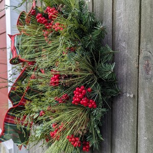 May include: A green and red plaid ribbon is tied around a wreath made of pine branches and red berries. The wreath is hanging on a weathered wooden door.