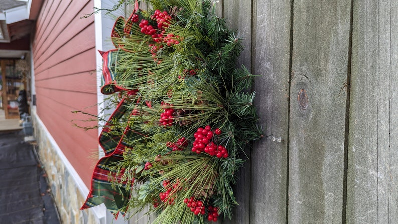 May include: A green and red plaid ribbon is tied around a wreath made of pine branches and red berries. The wreath is hanging on a weathered wooden door.