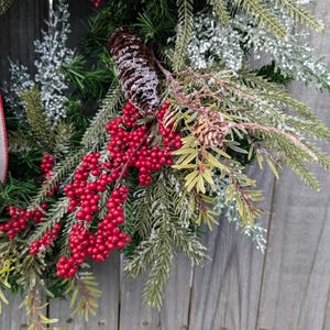 May include: A close-up of a Christmas wreath with red berries, pinecones, and evergreen branches. The wreath has a burlap ribbon with a red and white pattern. The background is a weathered wooden surface.