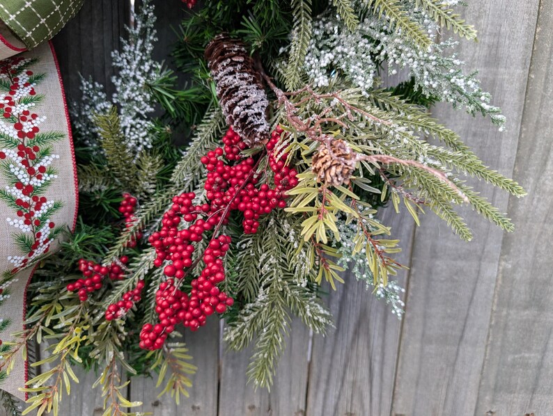 May include: A close-up of a Christmas wreath with red berries, pinecones, and evergreen branches. The wreath has a burlap ribbon with a red and white pattern. The background is a weathered wooden surface.