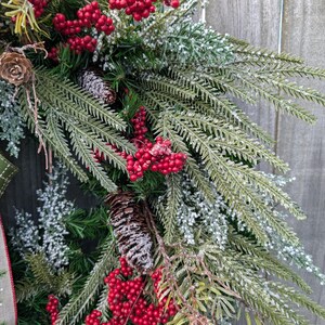 May include: A close-up of a Christmas wreath with evergreen branches, red berries, and pinecones. The wreath has a decorative ribbon with a green and white pattern. The background is a weathered wooden surface.
