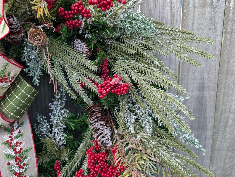 May include: A close-up of a Christmas wreath with evergreen branches, red berries, and pinecones. The wreath has a decorative ribbon with a green and white pattern. The background is a weathered wooden surface.