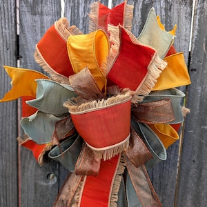 May include: A decorative bow constructed from multiple layers of wired ribbon in shades of burnt orange, mustard yellow, sage green, and metallic bronze. The ribbon edges feature a frayed, textured fringe detail throughout the entire arrangement.