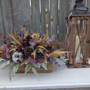 May include: A rustic wooden lantern and a floral centerpiece with a fall theme. The centerpiece features a wooden box filled with faux succulents, pinecones, berries, and dried foliage in shades of brown, purple, and green. Gold candle holders are also visible.