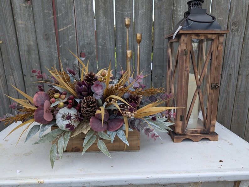 May include: A rustic wooden lantern and a floral centerpiece with a fall theme. The centerpiece features a wooden box filled with faux succulents, pinecones, berries, and dried foliage in shades of brown, purple, and green. Gold candle holders are also visible.