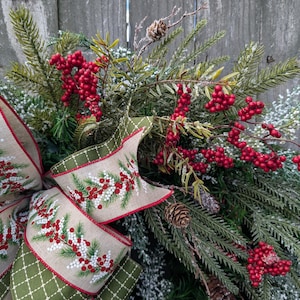 May include: A festive Christmas wreath featuring evergreen branches, red berries, and pinecones. A large bow with a tan background and a green diamond pattern edge is adorned with sprigs of red berries and white accents. The wreath is set against a weathered wooden background.