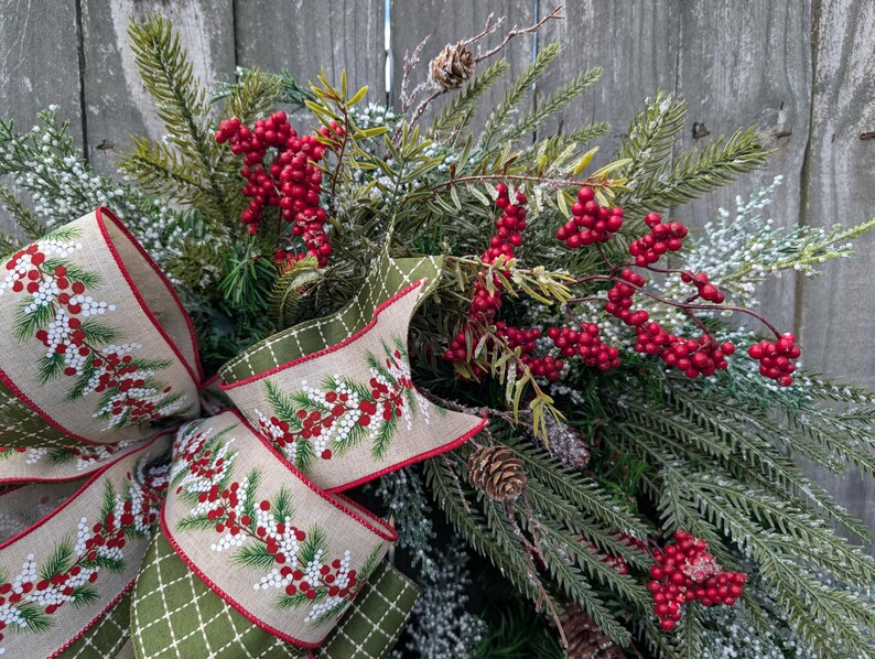 May include: A festive Christmas wreath featuring evergreen branches, red berries, and pinecones. A large bow with a tan background and a green diamond pattern edge is adorned with sprigs of red berries and white accents. The wreath is set against a weathered wooden background.