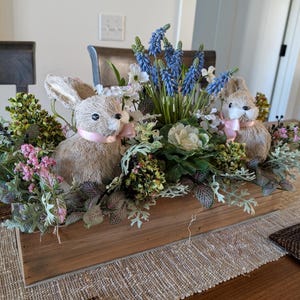 May include: A wooden box centerpiece with a variety of greenery, pink flowers, and two brown bunny figurines with pink ribbons. The centerpiece is on a brown table with a woven placemat.
