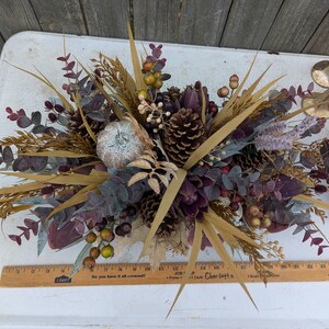 May include: A fall-themed floral arrangement featuring a small, silver pumpkin, pinecones, and various dried plants and berries. The arrangement includes eucalyptus leaves, wheat stalks, and purple accents. A wooden ruler is visible at the bottom.