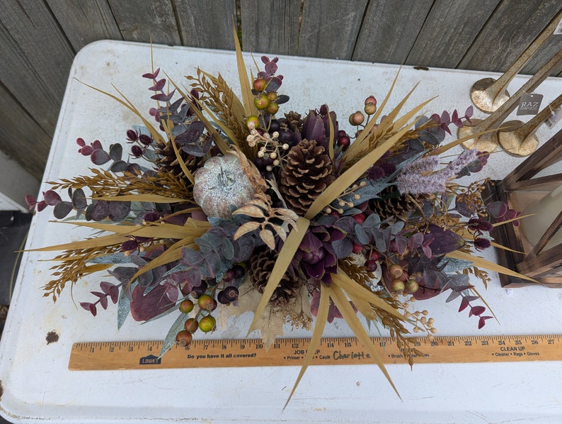 May include: A fall-themed floral arrangement featuring a small, silver pumpkin, pinecones, and various dried plants and berries. The arrangement includes eucalyptus leaves, wheat stalks, and purple accents. A wooden ruler is visible at the bottom.