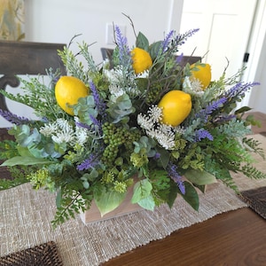 May include: A decorative centerpiece featuring bright yellow lemons nestled among faux greenery, including lavender and eucalyptus. The arrangement is set in a light wooden rectangular box, creating a fresh and vibrant display for a table or countertop.