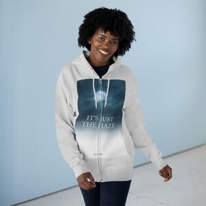 A light grey zip-up hoodie featuring a graphic of a full moon in a cloudy sky. The text "IT'S JUST THE HAZE" is printed on the graphic. The hoodie has a full zip and a hood.