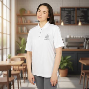 A woman stands indoors in a cafe wearing a white short-sleeve polo shirt with a small book stack graphic and text on the left chest.