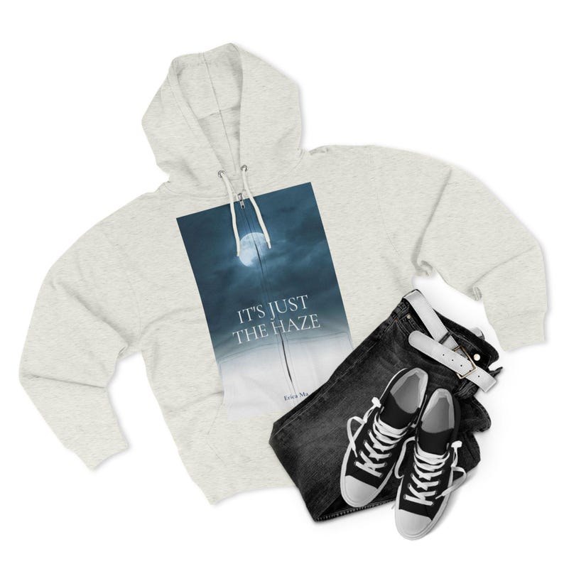 A light grey hoodie featuring a graphic of a moonlit sky with the text "IT'S JUST THE HAZE." The image also shows black jeans, a white belt, and black and white trainers.