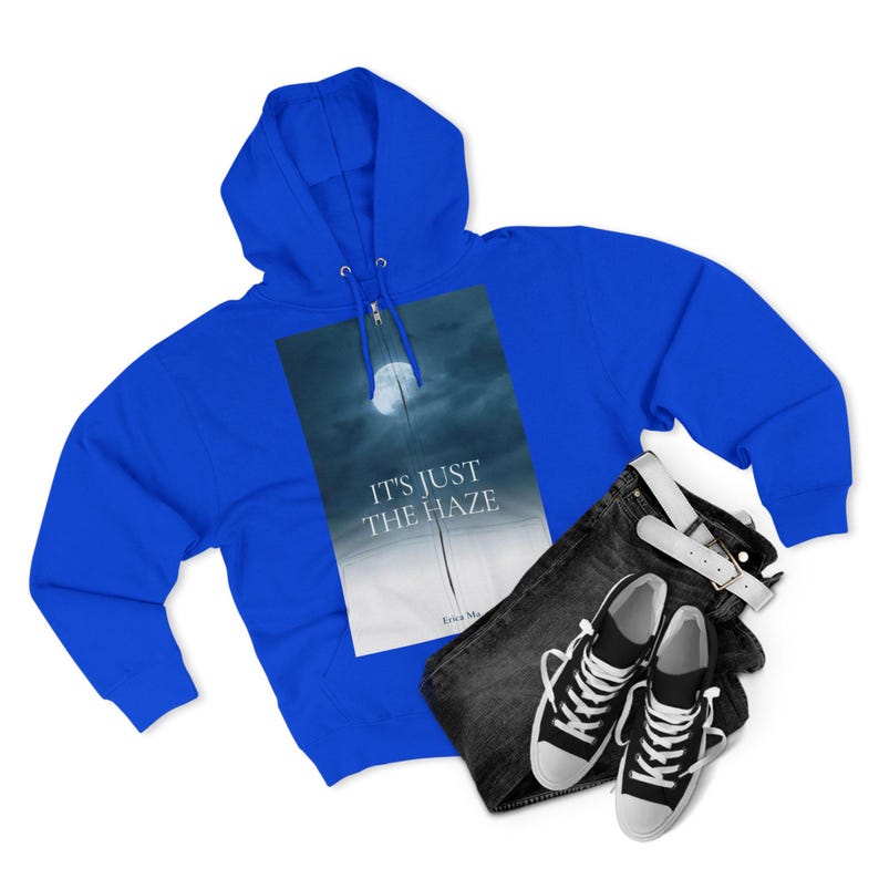 A vibrant blue hoodie featuring the text "IT'S JUST THE HAZE". The hoodie is displayed with black jeans, a white belt, and black and white trainers. The design includes a moon and a cloudy sky.