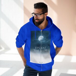 Royal blue zip-up hoodie featuring a full moon graphic in a cloudy sky. The text "IT'S JUST THE HAZE" is printed below the moon. The hoodie has a drawstring hood and a front zipper.