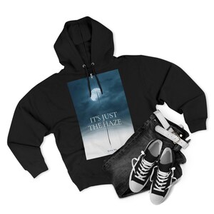 Black hooded sweatshirt with a full-length zip, displaying a graphic of a moon in a cloudy sky with the text "IT'S JUST THE HAZE." The sweatshirt is styled with dark denim jeans, a white belt, and black and white trainers.