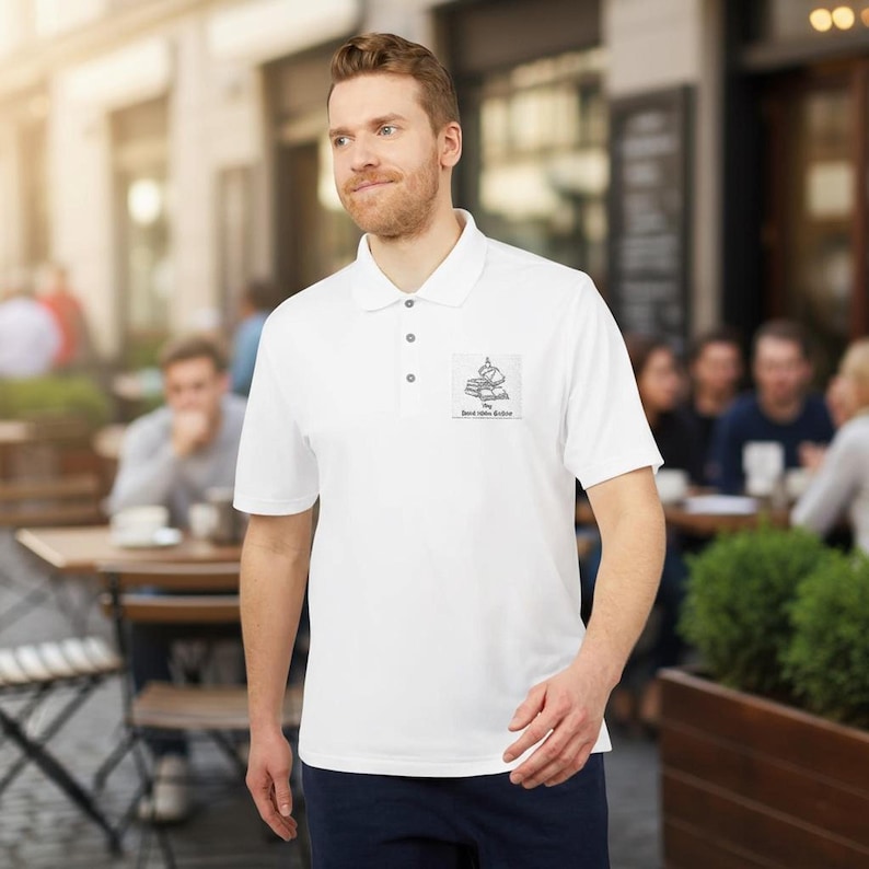 A man stands outdoors near a street cafe wearing a white short-sleeve polo shirt featuring a small book stack graphic on the chest.