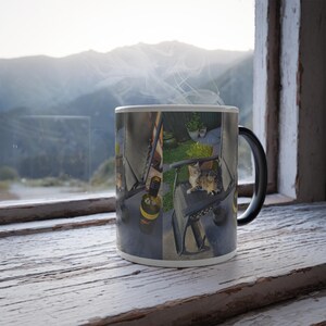May include: A black coffee mug featuring a wrap-around image of a cat on a chair, a wine bottle, and a mountain view. Steam rises from the mug. The mug is placed on a white, weathered wooden surface, suggesting a rustic setting.
