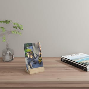 May include: A photograph of a cat sitting on a chair, displayed in a wooden photo holder. A bottle of wine and a small vase with greenery are also visible. A stack of magazines is on the right side of the image. The photo is on a wooden table.