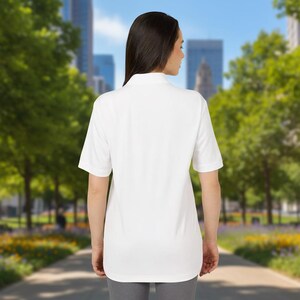 A woman stands outdoors with her back to the camera, wearing a plain white short-sleeve polo shirt and grey pants.